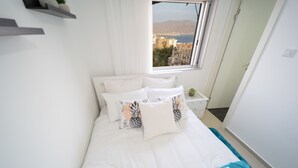 A neatly made bed with white bedding and decorative pillows, a window with a view of buildings and mountains, and a small bedside table with a plant. - YalaRent SeaView 3BR APT - Families only (Eilat)
