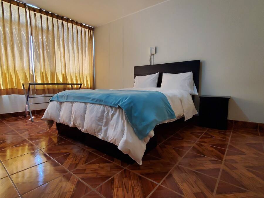 Een slaapkamer met een bed, nachtkastje, raam met gordijnen en een betegelde vloer.