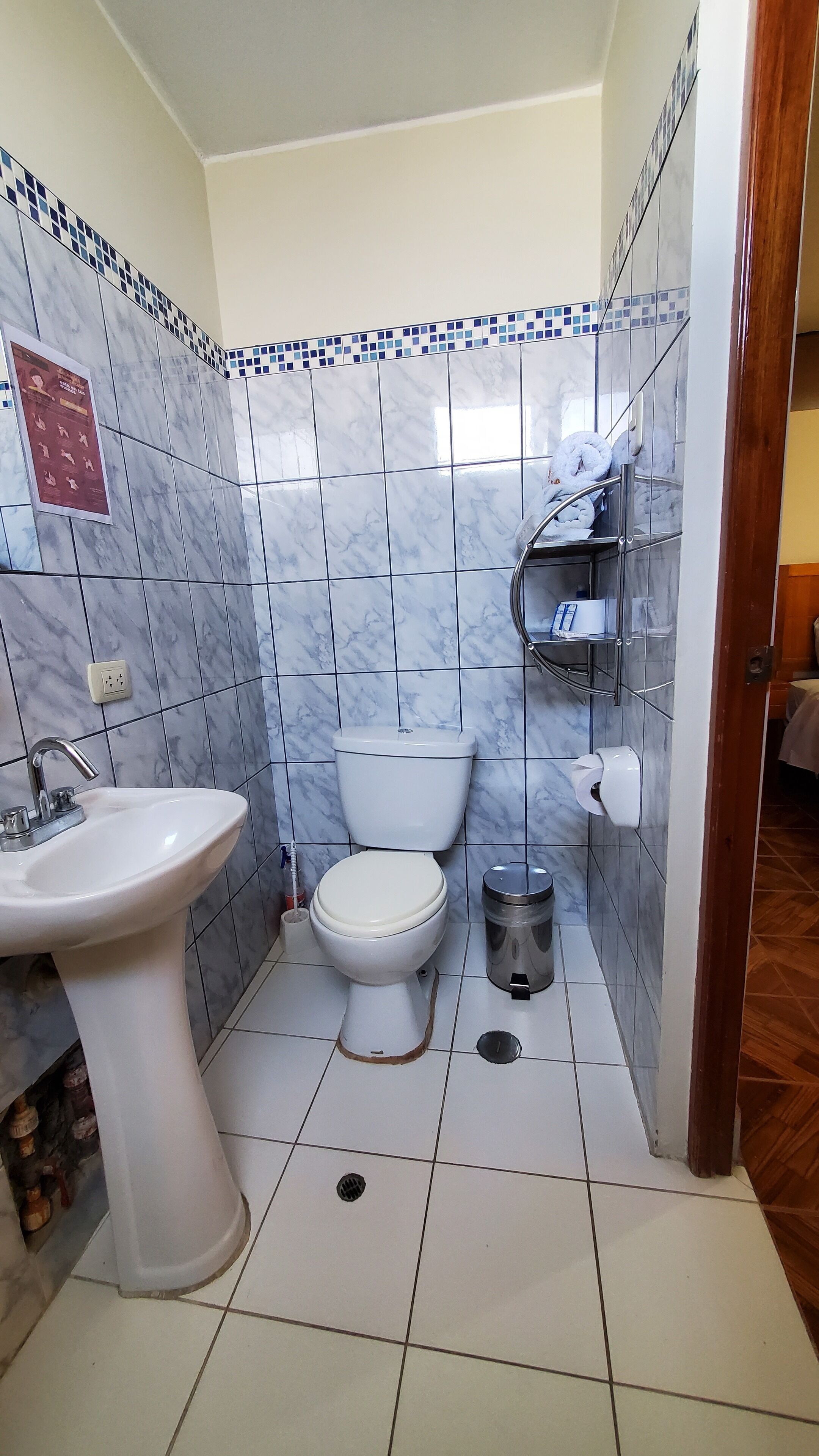Un baño con lavamanos, inodoro y cubo de basura blancos. Cuenta con un revestimiento de azulejos y un estante pequeño con artículos de tocador.