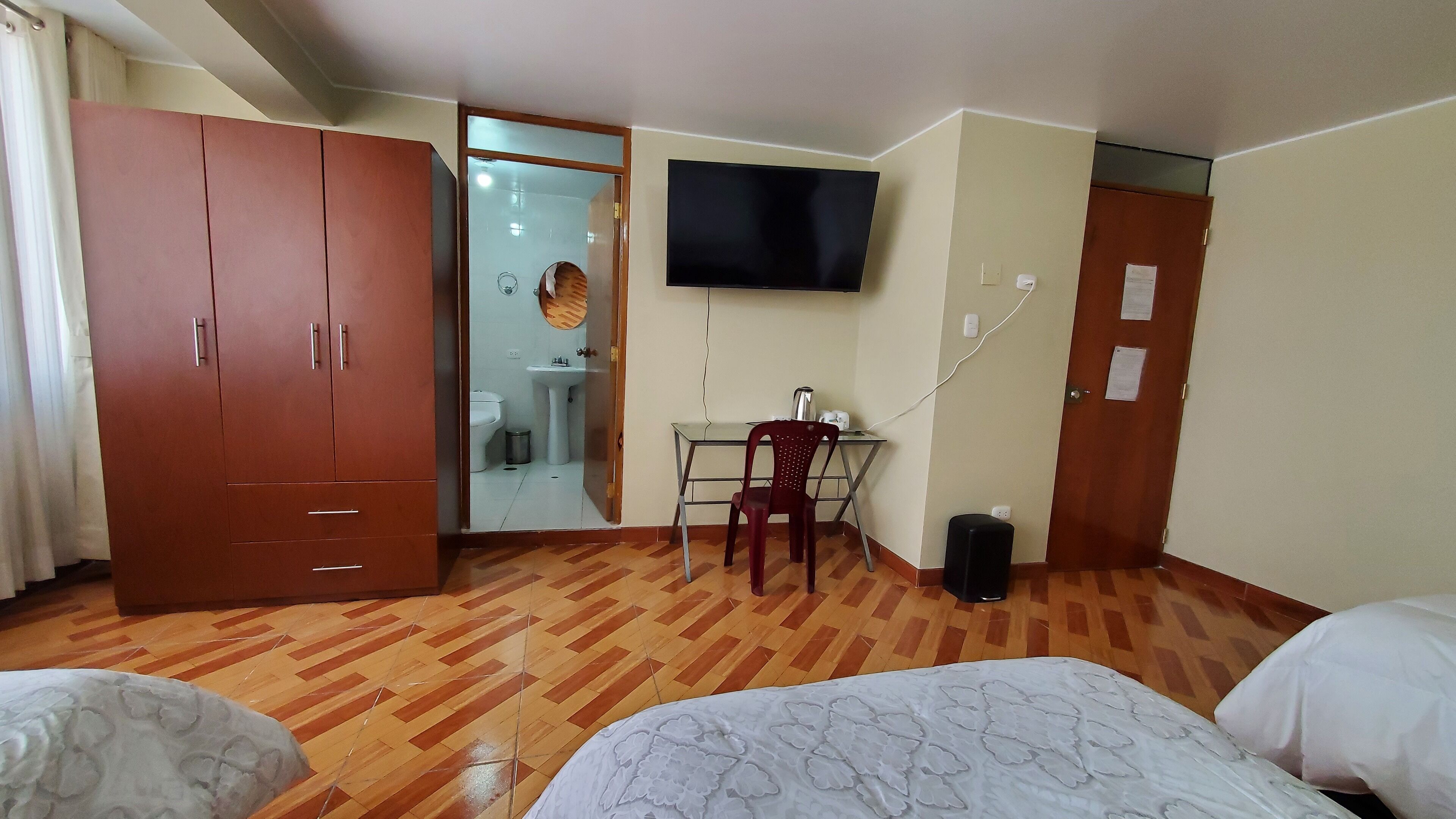 Ein Hotelzimmer mit Holzschrank, Schreibtisch mit Stuhl, Wandfernseher und einem Bett mit Blumenmuster-Bettdecke.