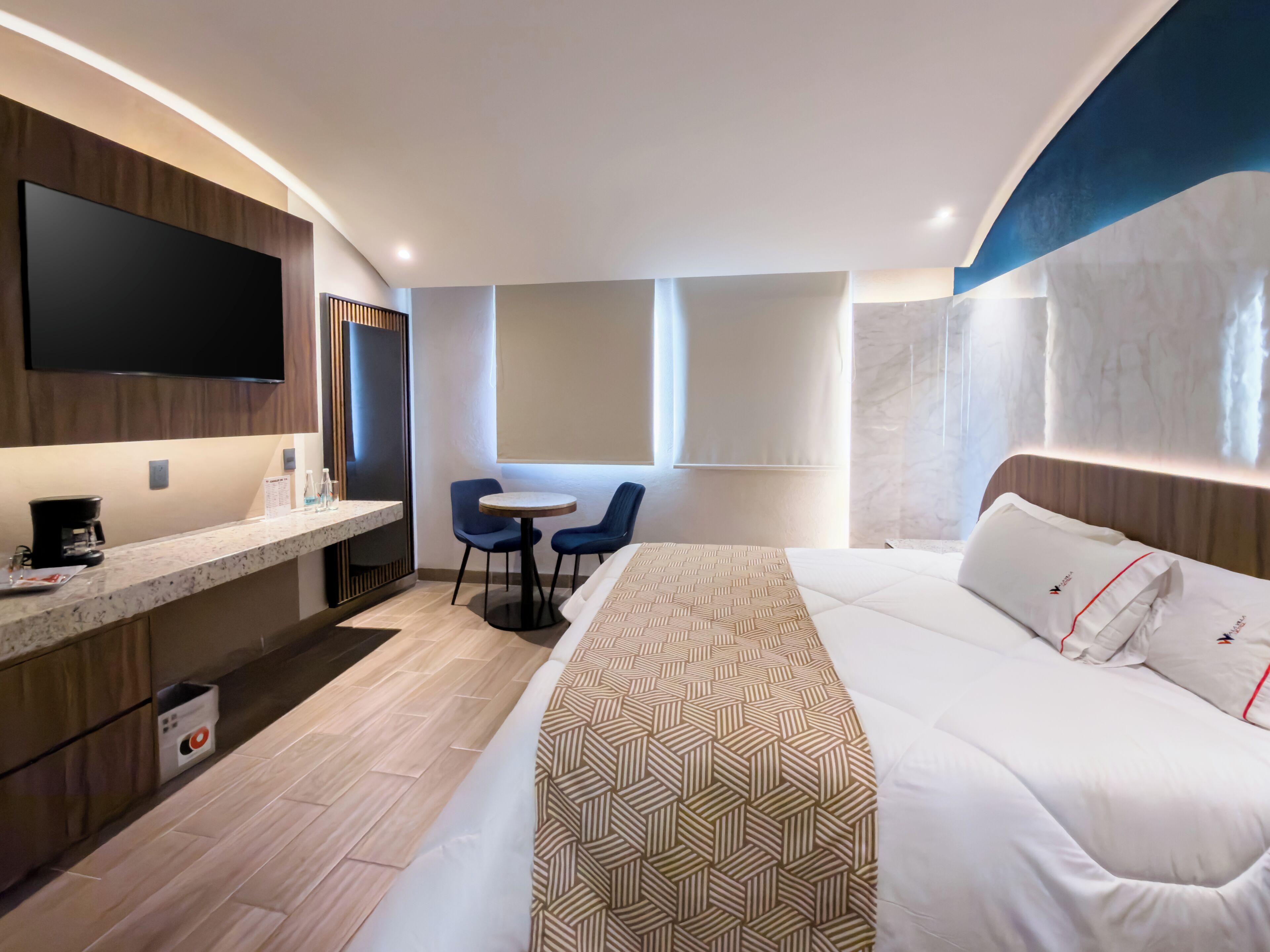 Una habitación de hotel moderna con una cama grande, un televisor de pantalla plana montado en la pared, una mesita con sillas y una ventana con cortinas.