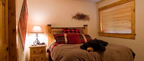 A cozy bedroom with a wooden bed, a nightstand with a lamp, a window with blinds, and a decorative wall piece.