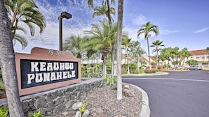 Ein Schild mit der Adresse „78 7070 Keanu Punahole“ und Blick auf eine Straße mit Palmen und einem Gebäude im Hintergrund.