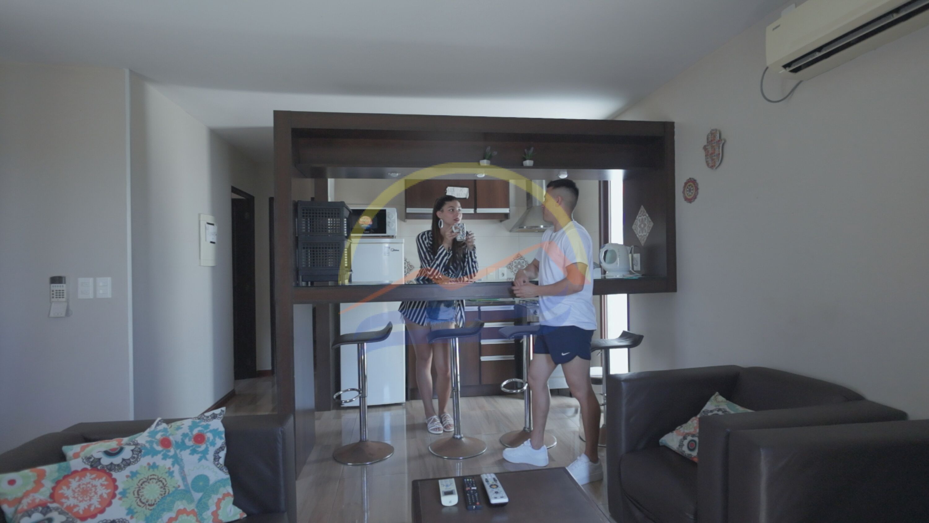 Una sala de estar con un sofá, una mesa de centro con controles remotos y una cocina al fondo.