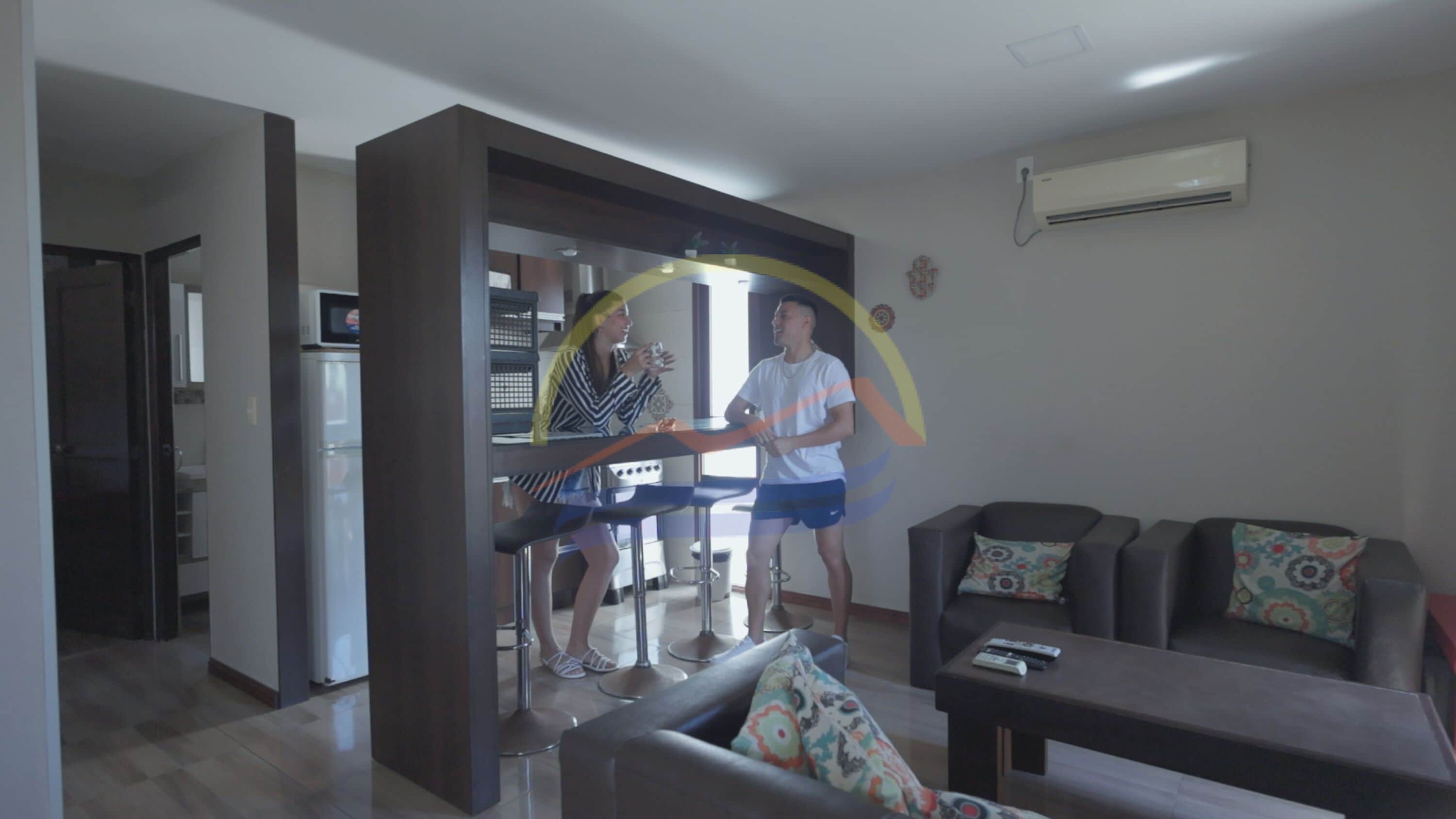 Una sala moderna con un sofá, una mesa de centro y una televisión. Se observa una cocina al fondo, con un refrigerador y un microondas.
