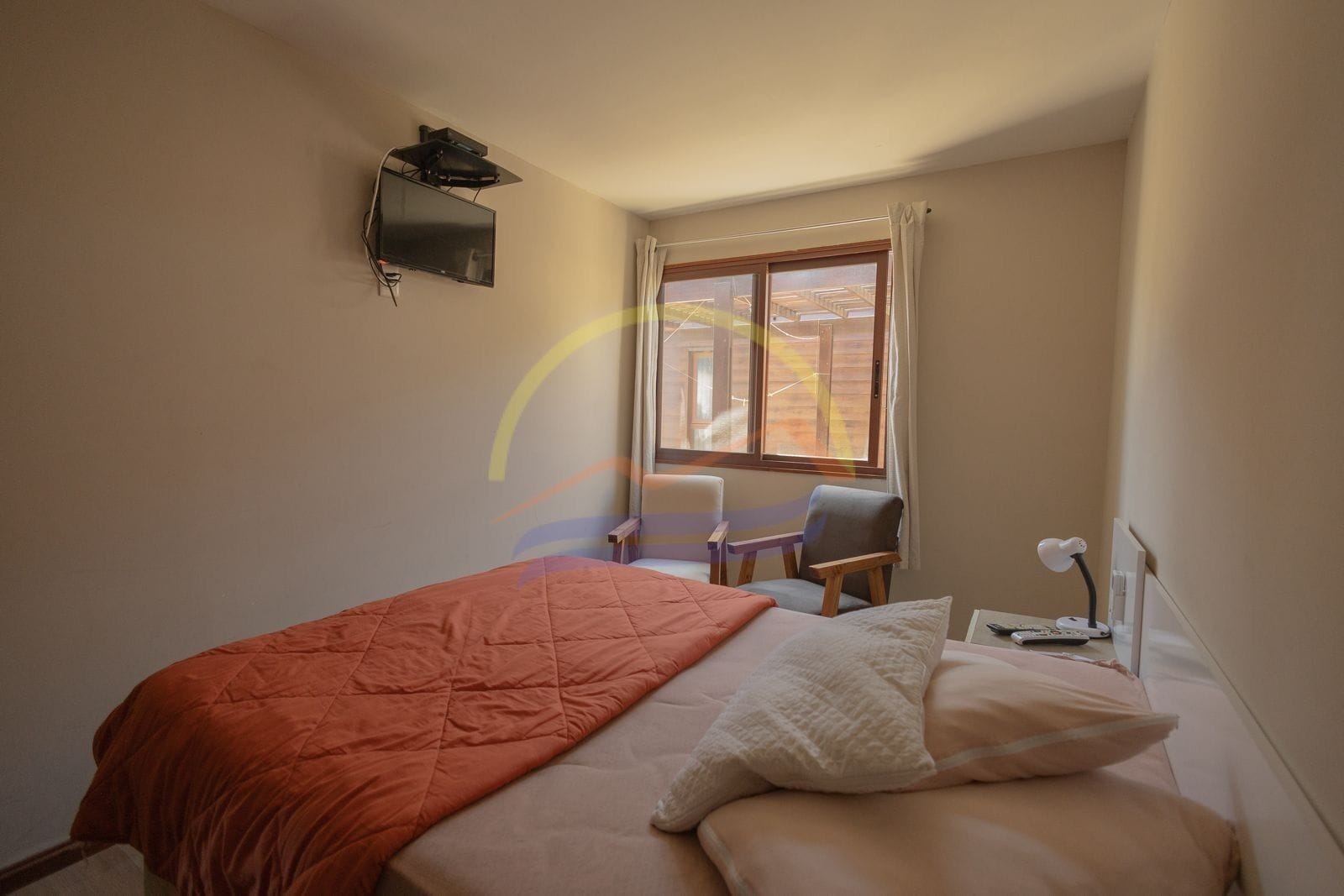 A bedroom with a bed, a chair, a desk, a TV, and a window with curtains.