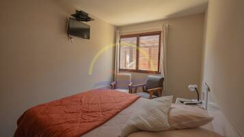 A bedroom with a bed, a chair, a desk, a TV, and a window with curtains.