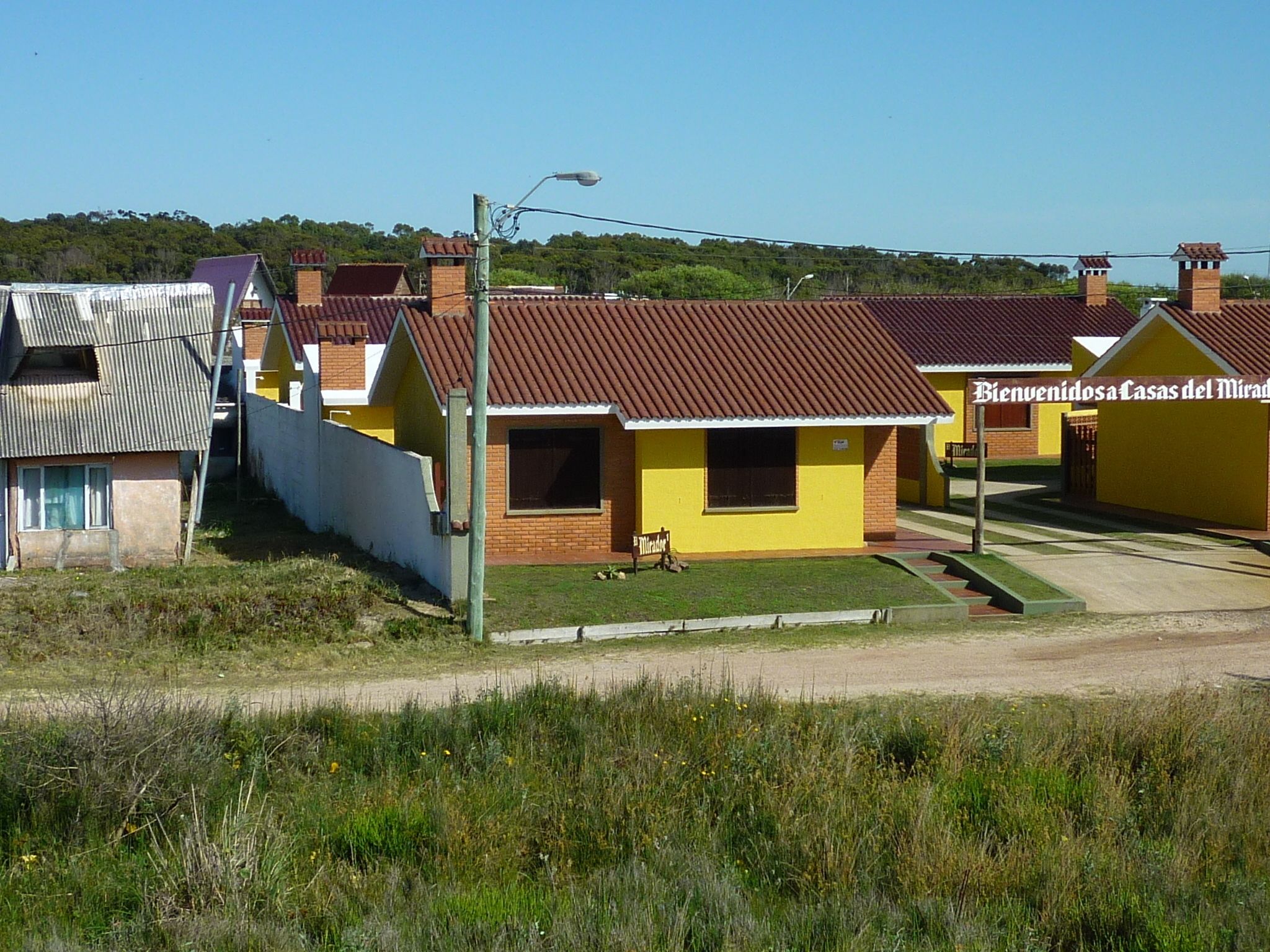 A row of yellow houses with brown roofs, each with a small front yard and a sign that reads "Bienvenidos Casas del Mirador".