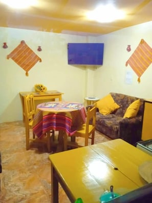 A small, cozy room with a round table covered with a colorful tablecloth, a sofa with yellow cushions, and a television mounted on the wall. - BiosWild (Cusco)