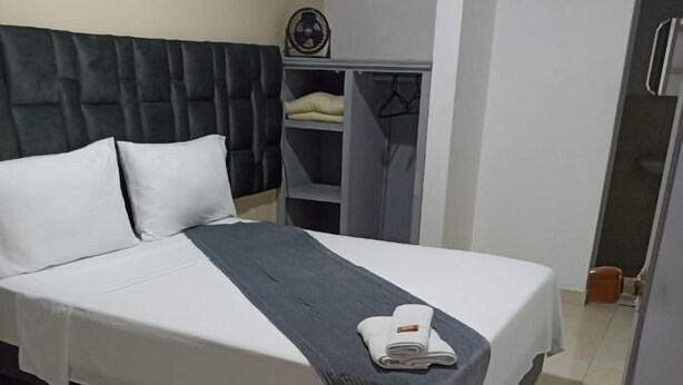 A neatly made bed with white and gray linens, a dark headboard, and a small fan on a shelf above. - Hotel Empresarial  (Pereira)