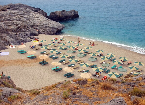 A beach with numerous umbrellas and lounge chairs, rocky terrain, and a view of the sea.