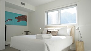 A bedroom with a large bed, a window with a mountain view, a painting on the wall, and a bedside lamp. - Basecamp Resorts Canmore, Basecamp Bargain Suite (Canmore)