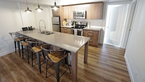 A modern kitchen with a central island, bar stools, stainless steel appliances, and wooden flooring. - Basecamp Resorts Canmore, Basecamp Bargain Suite (Canmore)