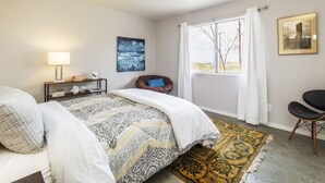 Una camera da letto con un letto, una ventola a soffitto, una finestra con tende, un comodino con lampada, una sedia e opere d'arte alle pareti.