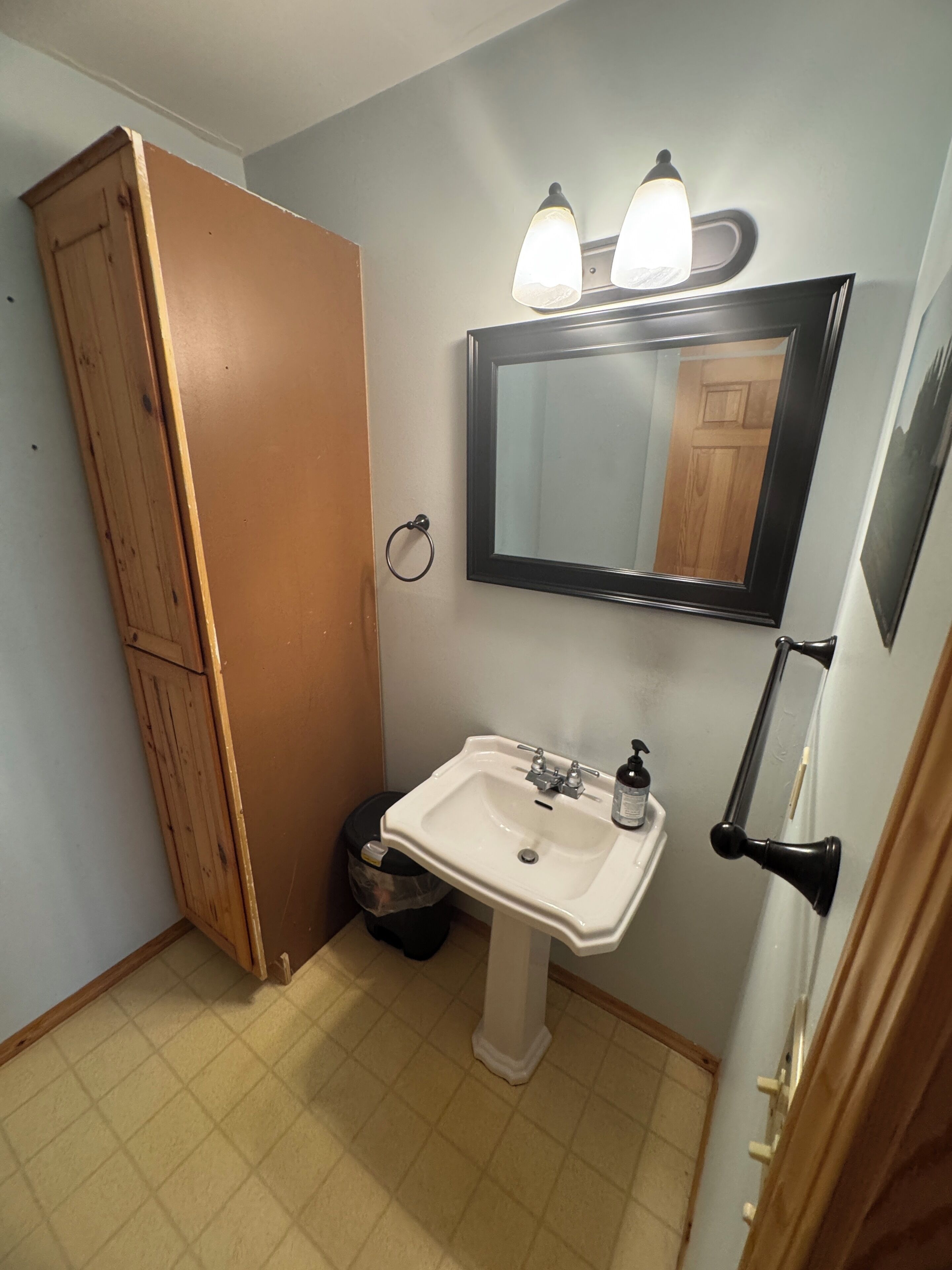 Une salle de bain avec un meuble en bois, un lavabo blanc avec un miroir au-dessus, et une poubelle.