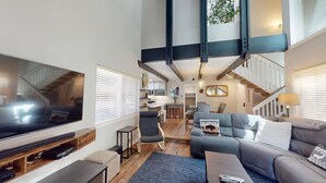 A spacious living area with a large flat-screen TV, comfortable seating, a staircase, and a kitchen in the background.