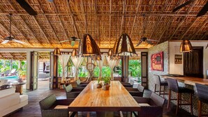 A spacious indoor area with a wooden table and wicker chairs, a thatched roof ceiling, and a view of greenery outside. - Unique beach house, luxury experience (Puerto Escondido)