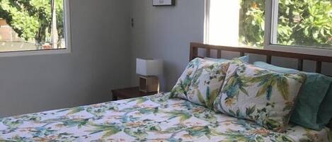A bedroom with a bed, a nightstand, a wall-mounted air conditioner, and large windows with a view of greenery.