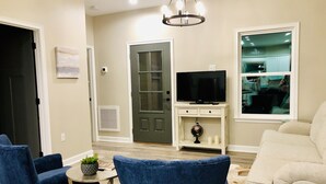 Ein Wohnzimmer mit Fernseher auf einem weißen Schrank, einem blauen Sessel, einem beigen Sofa und einem kleinen Tisch mit einer Pflanze.