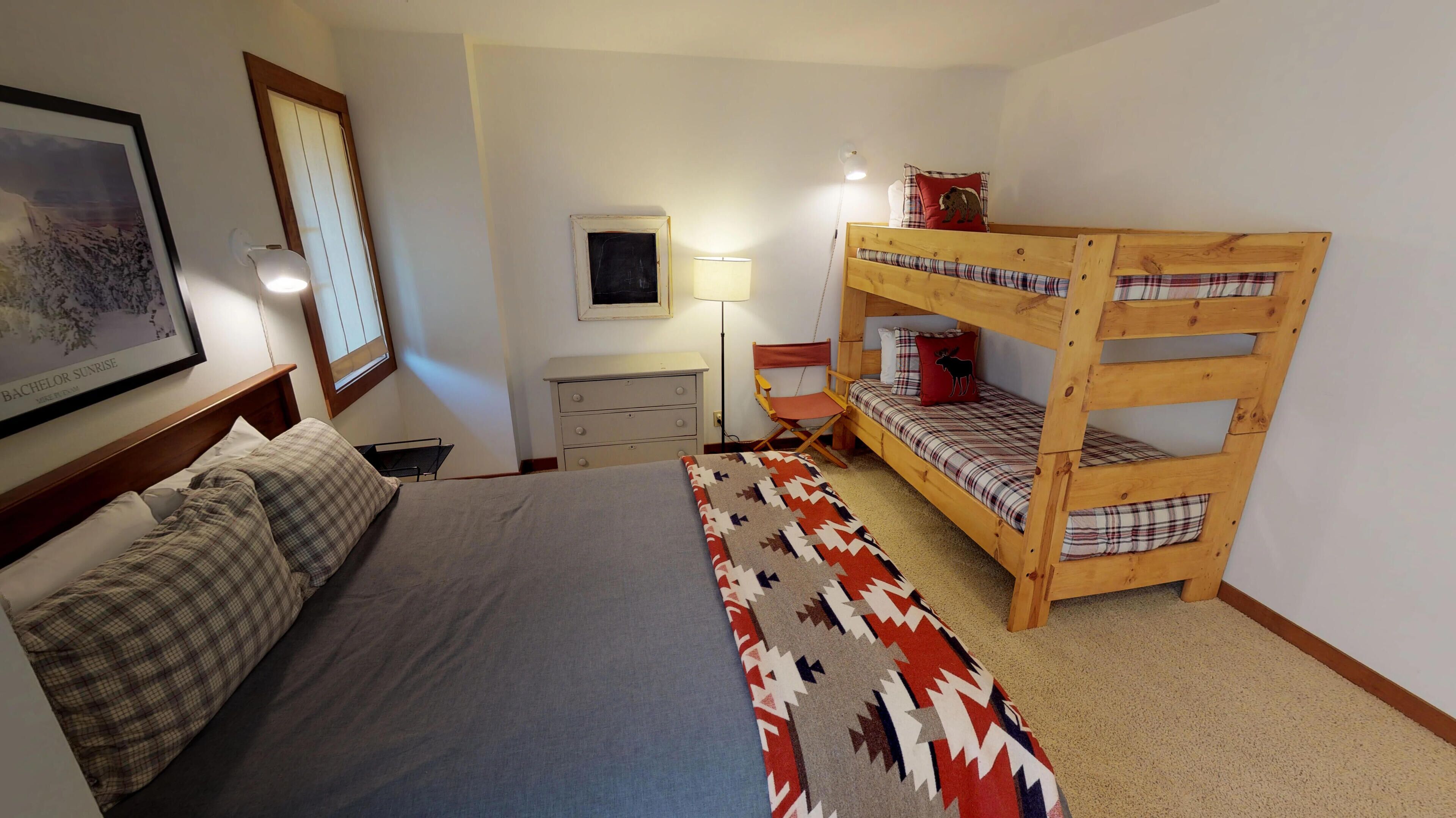 A bedroom with a bunk bed, a single bed, a dresser, a chair, a lamp, and a framed picture on the wall.