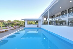 A modern pool area with lounge chairs and umbrellas, a glass railing, and a clear view of the surrounding greenery. - Evita Mare (Rhodes)