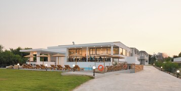 Un edificio moderno con zona de piscina, tumbonas, sombrillas y mobiliario de exterior.