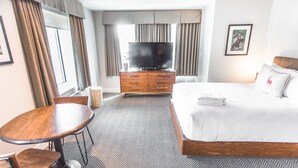 A hotel room with a large bed, a TV, a wooden dresser, a small round table, and a chair. - Basecamp Lodge I Micro-Suite Apartment (Canmore)