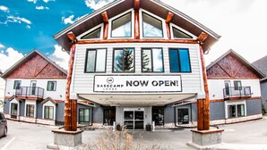 A lodge with a prominent "NOW OPEN" sign, featuring a modern architectural design with large windows and a stone foundation. - Basecamp Lodge I Micro-Suite Apartment (Canmore)
