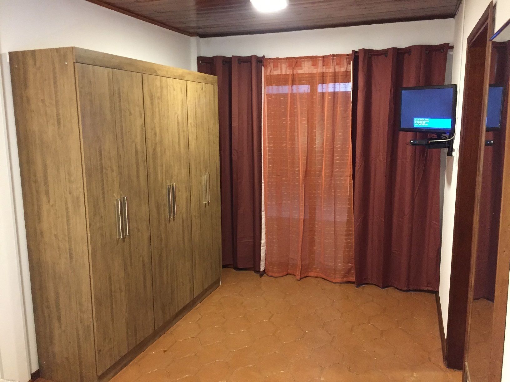 A room with a wooden wardrobe, a television on a stand, and a window with curtains.