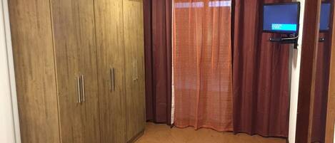 A room with a wooden wardrobe, a television on a stand, and a window with curtains.