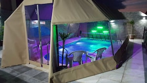 A covered outdoor swimming pool with a glass enclosure, surrounded by chairs and a small building.