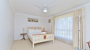 Ein Schlafzimmer mit einem Bett, einem Deckenventilator, einem Fenster mit Vorhängen, einem Kleiderständer und einem kleinen Tisch mit einer Lampe.