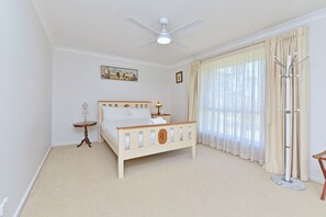 A bedroom with a bed, a ceiling fan, a window with curtains, a coat rack, and a small table with a lamp.