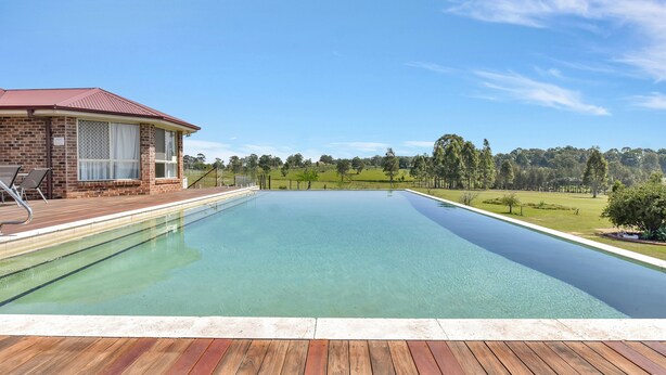 Ein rechteckiger Swimmingpool mit einer Holzterrasse im Vordergrund und ein Backsteinhaus mit rotem Dach im Hintergrund.