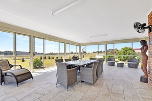 A spacious patio with a dining table and chairs, large windows offering a view of the countryside, and a brick wall with a fan.