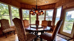 A wooden-paneled dining room with a round table and six patterned chairs, large windows with a view of trees, and a hanging light fixture. - Mountain Oasis Luxurious w/ 5 Star Amenities! (Bryson City)