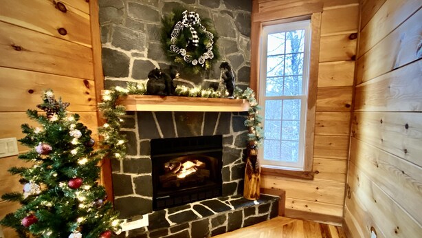 A cozy cabin interior with a stone fireplace, a Christmas tree, and a window with a view of trees. - Mountain Oasis Luxurious w/ 5 Star Amenities! (Bryson City)