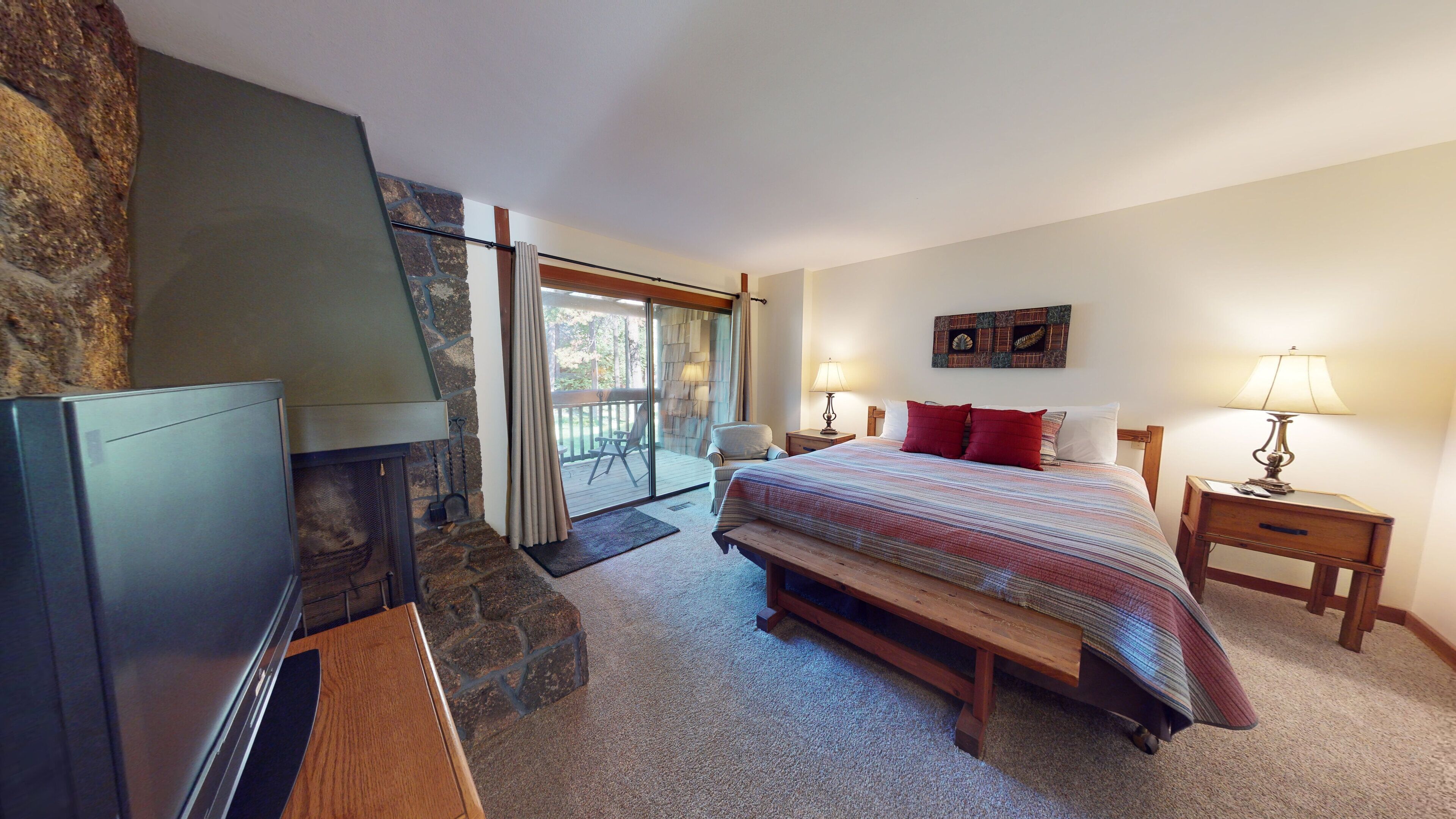 A bedroom with a stone fireplace, a bed with red pillows, a wooden bench, and a nightstand with a lamp.