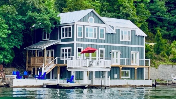 Una casa de dos pisos con un muelle, sillas azules y un paraguas rojo.