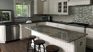Una cocina con armarios blancos, encimera de granito y salpicadero con un diseño de rombos. Hay tres taburetes de bar y una ventana con vistas a zonas verdes.