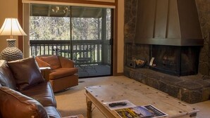 A cozy living room with a leather sofa, a coffee table with a remote control and magazine, a fireplace, and a large sliding glass door leading to an outdoor deck. - Cozy Sunriver Golf Condo with Access to Exclusive Golf (Sunriver)