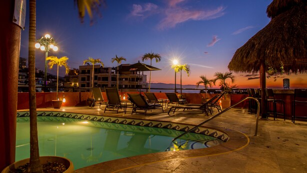 A pool area with lounge chairs, a thatched-roof structure, and a view of the sunset over the ocean. - Andales on the Bay Suites (Puerto Vallarta)
