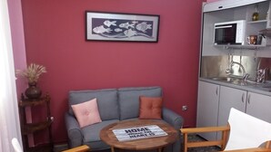 En hyggelig opholdsstue med en grå sofa, to træstole, et lille rundt bord med en avis, et indrammet kunstværk over sofaen, en mikroovn og en vask i baggrunden.