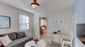 Una sala de estar con un sofá gris, una mesa de centro blanca con un jarrón de flores, un comedor con una mesa y sillas blancas, y una cocina visible a través de un umbral abierto.