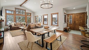 Un salón comedor amplio con una mesa de madera, bancos y una araña. Ventanas grandes ofrecen luz natural y vistas al exterior.