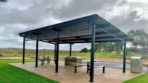 A covered outdoor seating area with benches and tables, surrounded by a grassy field and a metal fence.
