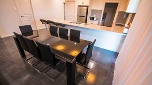 A modern dining area with a rectangular table and six chairs, a kitchen in the background with a refrigerator and microwave, and a door to the left.