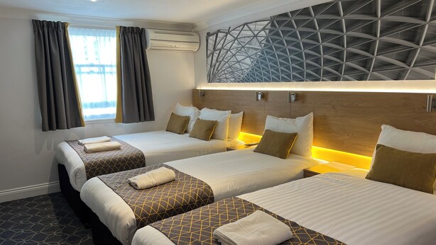 A hotel room with three beds, a modern ceiling design, and a large window with curtains. - The Suites- St Pancras Hotel Group (London)