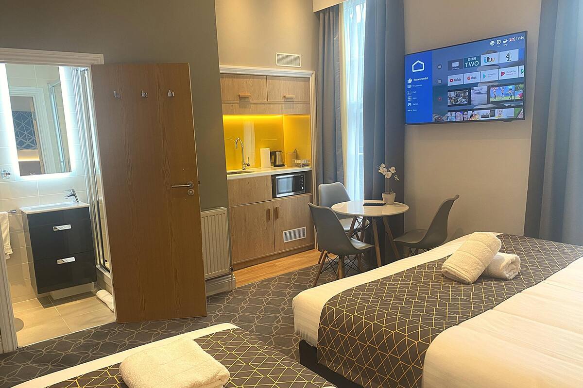 Camera d'albergo con due letti, angolo cottura, bagno e televisore che mostra un servizio di streaming.