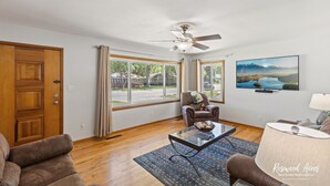 Uma sala de estar espaçosa com piso de madeira, um ventilador de teto, uma janela grande com cortinas, uma poltrona de couro marrom, uma mesa de centro de vidro e um quadro na parede.